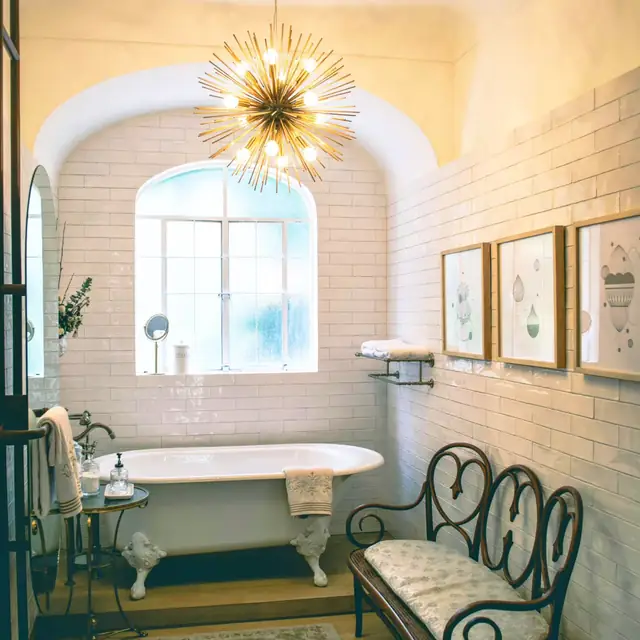 A clawfoot tub under an arched window in a white-tiled bathroom, lit by a starburst chandelier.