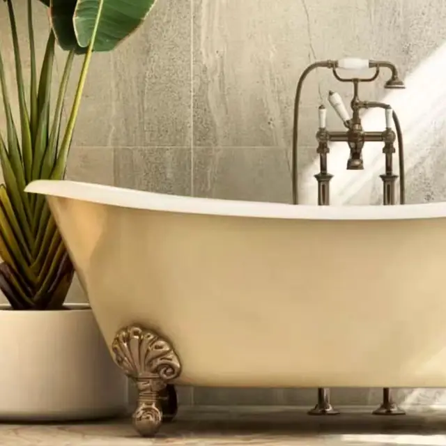 A cream clawfoot tub beside a potted plant, with an aged brass tub filler against a stone-tiled wall.