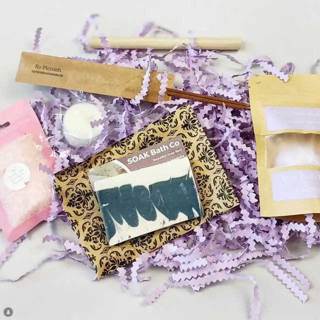 Gift box contents on lilac paper shred: a bar of marbled soap, pouches of bath salts and other small bath goods.