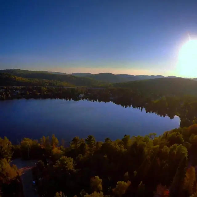 Aerial view of a lake and forested hills at sunset, the sun low over the far shore.